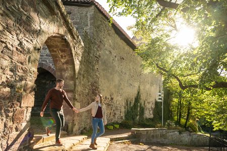 Ein Paar spaziert Hand in Hand an einer alten steinernen Mauer entlang, umgeben von grünen Bäumen.