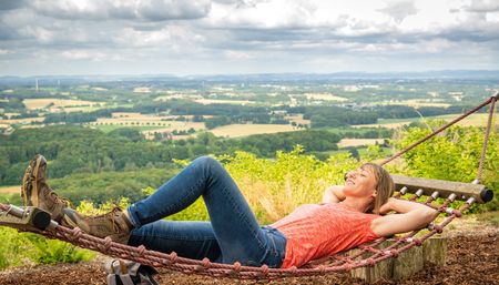 Frau entspannt sich in einer Hängematte mit weitem Blick auf grüne Landschaft und bewölkten Himmel.