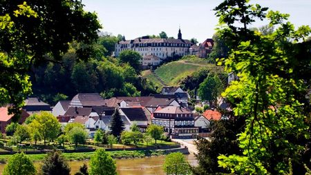 Blick auf die Abtei vom Heiligen Kreuz in Herstelle, umgeben von dichten Wäldern und einem Fluss.