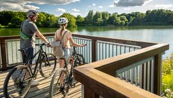 Ein Paar mit Helmen genießt auf einer Holzbrücke die Aussicht auf einen See inmitten grüner Natur.