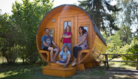 Familie sitzt lächelnd vor einer runden, hölzernen Campinghütte auf sonnigem, grünem Gelände.
