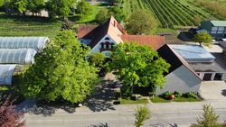Luftaufnahme des Obsthofs mit Gebäude, umgeben von Gärten und Gewächshäusern, bei sonnigem Wetter.
