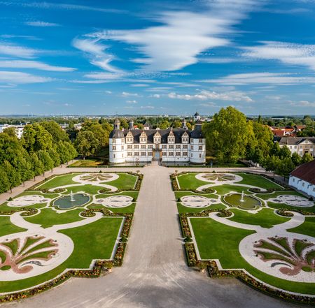 Barockschloss mit symmetrischem, kunstvollem Garten aus der Vogelperspektive unter klarem Himmel.