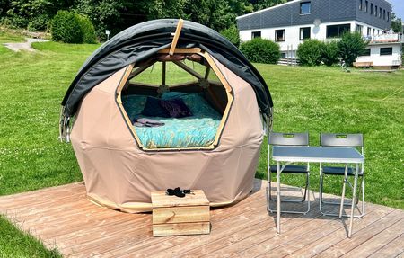 Beiges Sternkuppelzelt mit Campingstühlen und Tisch auf Rasen, im Hintergrund das Bootshaus.