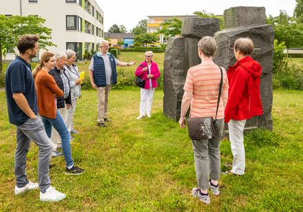 Eine Gruppe von Menschen betrachtet eine moderne Steinskulptur in einem parkähnlichen Gelände.
