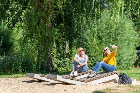 Zwei lachende Frauen entspannen sich bei Sonnenschein auf einer Liegebank in einem grünen Park.