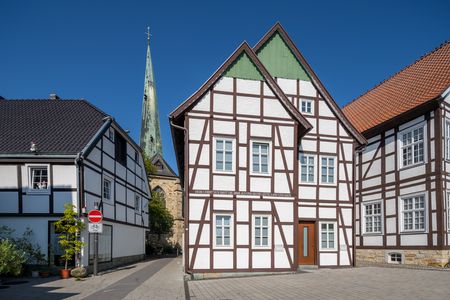 Fachwerkhäuser vor blauem Himmel mit Kirchturm im Hintergrund, gepflasterte Straße ohne Verkehr.