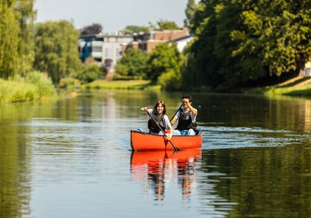 Zwei fröhliche Menschen paddeln in einem roten Kanu auf einem ruhigen, von Bäumen gesäumten Fluss.