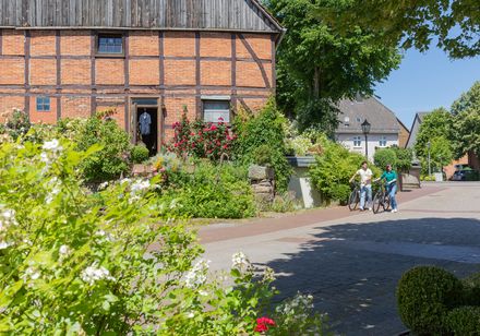 Fachwerkhaus in Marienmünster-Vörden mit rotem Ziegel, umgeben von blühenden Gärten; zwei Radfahrer fahren vorbei.