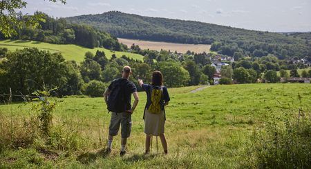 Zwei Wanderer betrachten eine grüne Hügellandschaft mit Wäldern und einem malerischen Dorf im Tal.