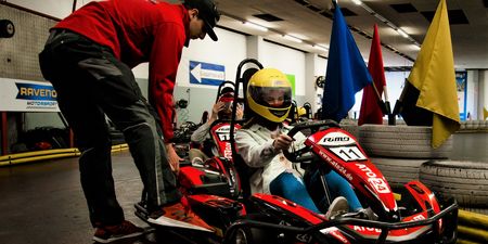 Ein Kind im gelben Helm sitzt in einem roten Go-Kart, betreut von einem Erwachsenen in einer Halle.