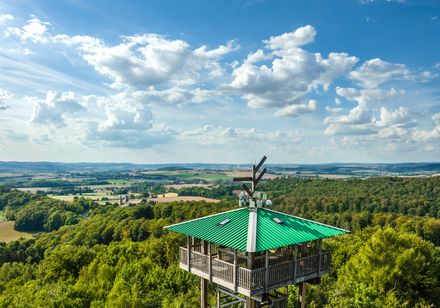 Holzturm mit grünem Dach (Hungerbergturm) über grüner Waldlandschaft, im Hintergrund weitläufiges Tal und blauer Himmel.