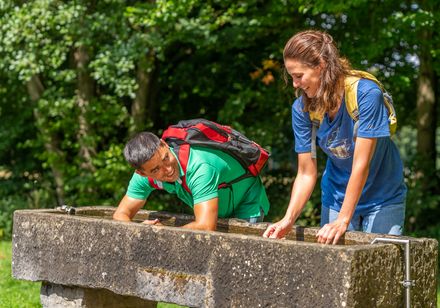 Zwei Menschen beugen sich über einen steinernen Brunnen im Grünen, lächelnd und bei sonnigem Wetter.