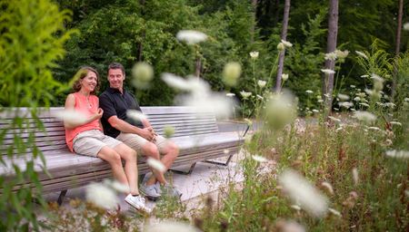 Zwei Personen sitzen lachend auf einer Parkbank im Grünen, umgeben von blühenden Wildblumen.