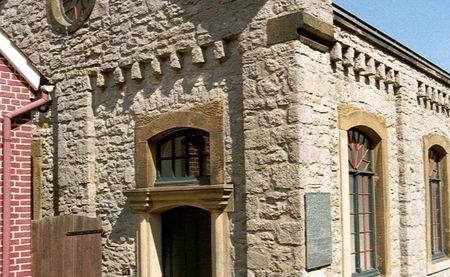 Kleine Synagoge aus hellem Stein mit Spitzgiebel, Rundfenstern und grüner Holztür, lateralansicht.