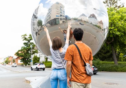 Zwei Menschen stehen vor einer großen, spiegelnden Kugel, die eine Straße und Gebäude reflektiert.