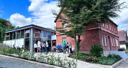 Das Bild zeigt ein renoviertes Backsteingebäude mit moderner Glasfassade und versammelten Personen.