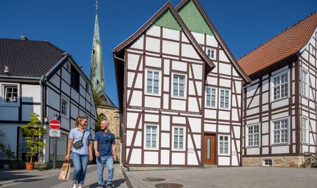 Fachwerkhäuser und ein Kirchturm in einem sonnigen, historischen Stadtzentrum mit zwei Passanten.
