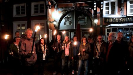 Menschen halten Fackeln vor einem beleuchteten, historischen Fachwerkgebäude in abendlicher Szenerie.