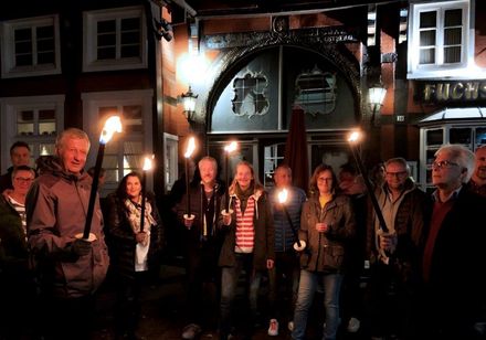 Menschen halten Fackeln vor einem beleuchteten, historischen Fachwerkgebäude in abendlicher Szenerie.