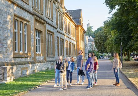 Gruppe von acht Personen betrachtet die historische Fassade eines alten Steingebäudes in sonniger Umgebung.