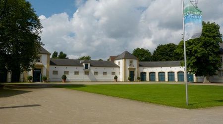 Heller Gebäudekomplex mit Torbögen und blauen Türen, flankiert von Rasenfläche und Fahne.